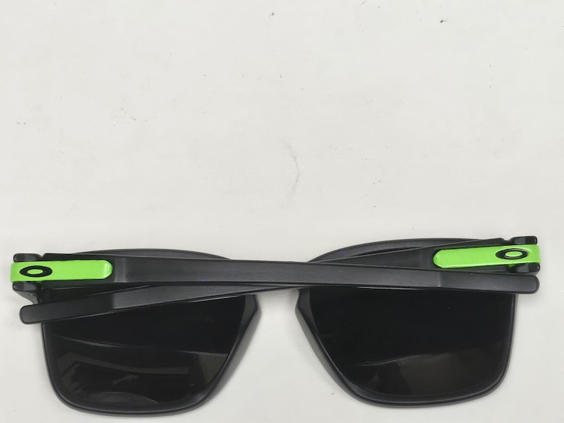 May include: Black sunglasses with green accents on the arms. The lenses are black and the frame is made of plastic.