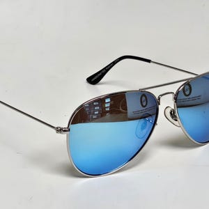 Puede incluir: Gafas de sol estilo aviador con montura plateada y lentes azules espejadas. Las patillas son negras y el texto "HANDCRAFTED" es visible en la patilla. Las lentes reflejan el entorno.