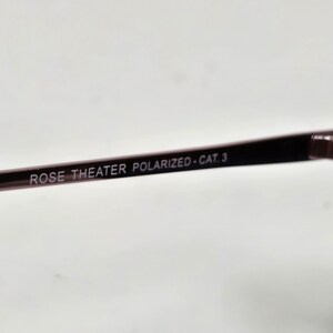 May include: A dark-colored eyeglass temple arm featuring the printed text ROSE THEATER POLARIZED - CAT. 3. The arm is thin and smooth, connecting to a larger frame component on the right side of the image, showcasing a sleek and functional design.