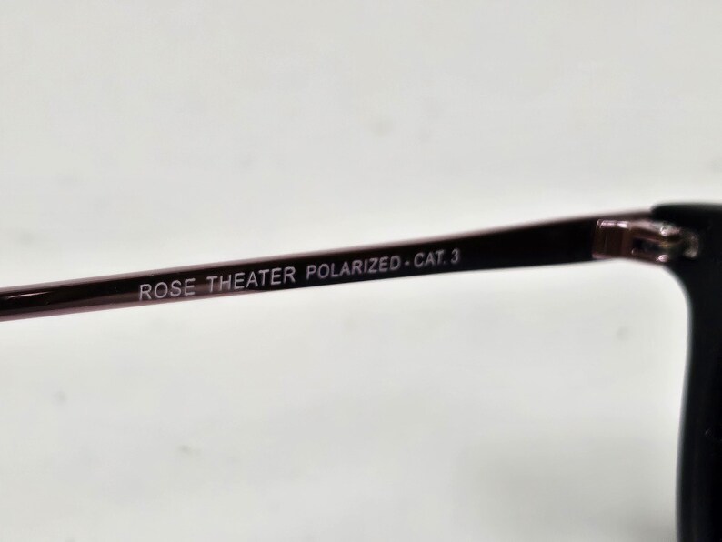 May include: A dark-colored eyeglass temple arm featuring the printed text ROSE THEATER POLARIZED - CAT. 3. The arm is thin and smooth, connecting to a larger frame component on the right side of the image, showcasing a sleek and functional design.