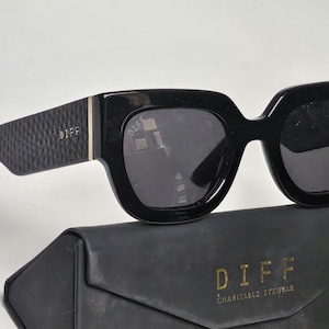 May include: Black square-framed sunglasses with dark lenses. The temples have a textured pattern and the brand name "DIFF" is visible. The sunglasses rest on a black case with the brand name and "CHARITABLE EYEWEAR" printed in gold.