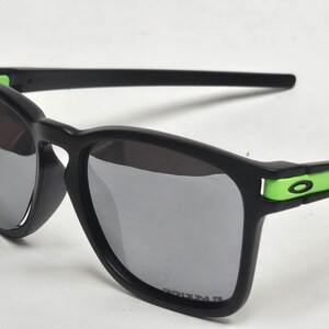 May include: Black sunglasses with green accents and gray lenses. The lenses are labeled "PRIZM".