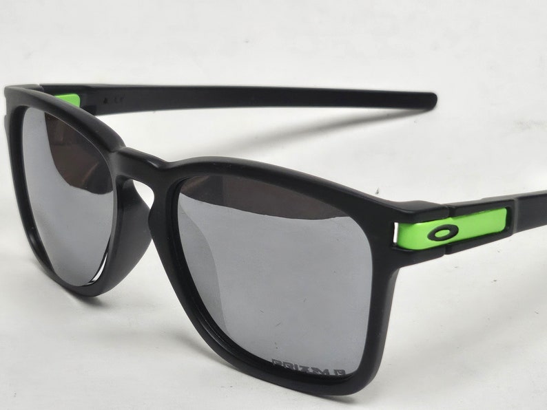 May include: Black sunglasses with green accents and gray lenses. The lenses are labeled "PRIZM".