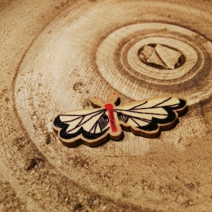 Può includere: Una farfalla di legno con ali bianche e nere e un centro rosso, seduta su una superficie di legno marrone.