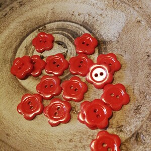 May include: A collection of red flower-shaped buttons with two white holes in the centre. The buttons are scattered on a wooden surface.
