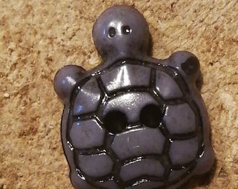 Może przedstawiać: Guzik ceramiczny w kolorze fioletowym w kształcie żółwia z czarnym obramowaniem. Guzik ma fakturowany pancerz i dwa małe otwory do szycia.