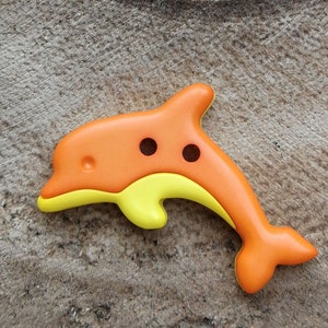 Peut inclure: Bouton en forme de dauphin orange et jaune, avec deux trous noirs pour les yeux. Le bouton a une forme arrondie et est posé sur un fond texturé de tons neutres. Le ventre du dauphin est jaune, et le reste du bouton est orange.