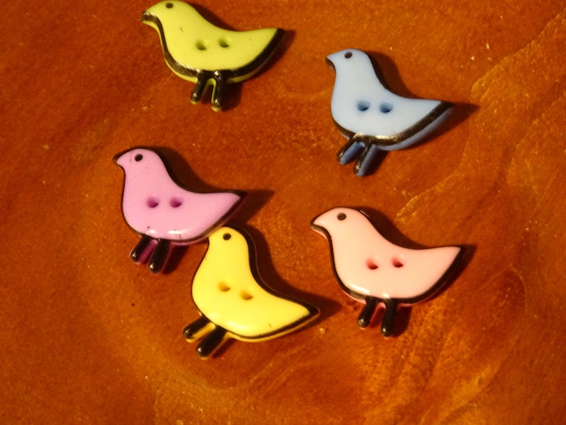 Pode incluir: Cinco bot&otilde;es de pl&aacute;stico coloridos em forma de p&aacute;ssaro com contornos pretos. Os bot&otilde;es s&atilde;o amarelos, rosa, azul claro e dois tons de rosa.