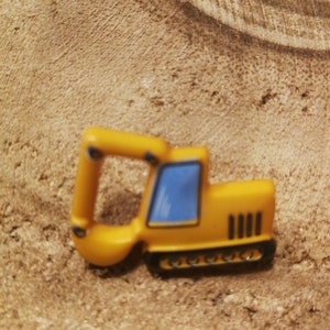 Może przedstawiać: Mała, żółta zabawka koparka z niebieskim oknem i czarnymi gąsienicami. Koparka ma ruchome ramię i jest umieszczona na teksturowanym, jasnobrązowym tle.