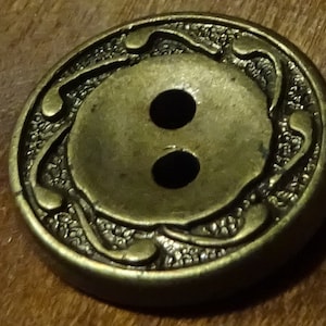 Peut inclure: Un bouton en métal doré avec un motif décoratif autour du bord. Le bouton a deux trous au centre.