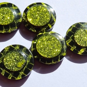 Könnte beinhalten: Fünf runde schwarze und grüne Knöpfe mit einem glitzernden grünen Zentrum. Die Knöpfe haben ein schwarz-gelbes Karomuster am Rand.