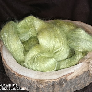 Pode incluir: Um novelo de fio verde claro numa tigela de madeira. O fio é macio e fofo, e é perfeito para tricotar ou fazer crochê.