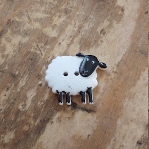 May include: A sheep-shaped button, white with a black head and legs, against a wooden background. The button has two holes. Suitable for sewing and craft projects, this button adds a charming touch.