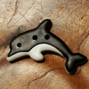 May include: A black and white dolphin-shaped button with three black dots for eyes. The button is made of plastic and has a smooth, glossy finish.