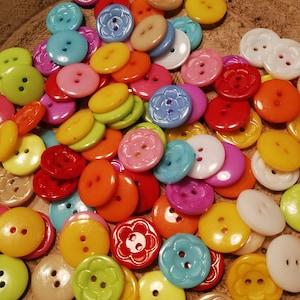 Puede incluir: Una colección de botones de plástico de colores en varias formas y tamaños, incluyendo botones redondos, con forma de flor y de dos agujeros.