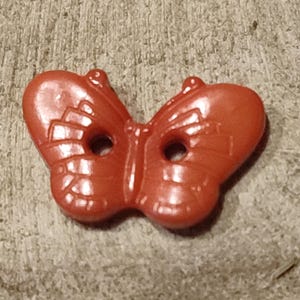 Könnte beinhalten: Ein roter, schmetterlingsförmiger Knopf mit zwei Löchern. Der Knopf hat detaillierte Flügelmuster und eine glänzende Oberfläche. Dieser dekorative Knopf eignet sich für Nähprojekte und Bastelarbeiten.