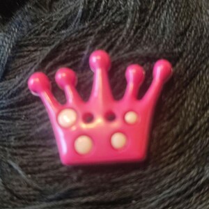 Könnte beinhalten: Ein leuchtend rosa kronenförmiges Ornament mit weißen kreisförmigen Akzenten. Die Krone hat fünf abgerundete Spitzen und drei kleine Löcher. Das Objekt befindet sich vor einem dunklen, strukturierten Hintergrund.