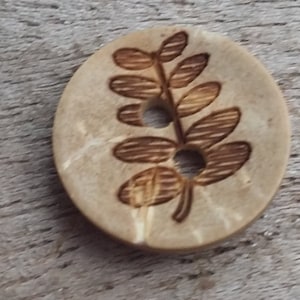 May include: A circular, light brown wooden button featuring a leaf design etched in darker brown. The button has a central hole for sewing and a natural wood grain appearance. Suitable for sewing projects.