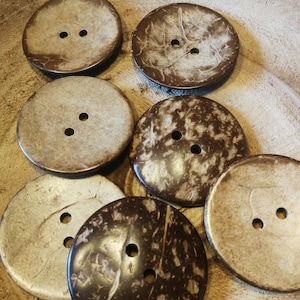 Könnte beinhalten: Sechs braune Kokosnuss-Knöpfe aus Kokosnussschale, jeder mit zwei Löchern. Die Knöpfe haben ein natürliches, rustikales Aussehen und sind perfekt, um Ihren Bastelarbeiten einen Hauch von Natur zu verleihen.