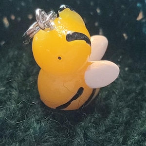Könnte beinhalten: Ein gelber und schwarzer Bienenanhänger mit weißen Flügeln. Die Biene hat eine silberne Schlaufe zum Anbringen an einer Halskette oder einem Armband.