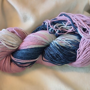 Könnte beinhalten: Ein Knäuel Garn in Rosa, Blau und Weiß. Das Garn ist fest gewickelt und wirkt weich. Die Farben sind ineinander übergehend, wodurch ein Farbverlauf entsteht. Das Garn wird wahrscheinlich für Strick- oder Häkelprojekte verwendet.