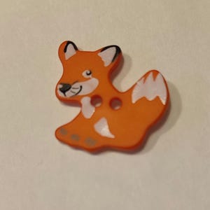 Könnte beinhalten: Orangener Plastikknopf mit einem weißen Fuchsdesign. Der Fuchs hat schwarze Augen und eine schwarze Nase.