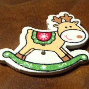 Op de afbeelding: Een houten kerstversiering in de vorm van een rendier-schommelpaard. Het rendier is bruin met een rode en groene hulstkrans om zijn nek. Het schommelpaard is wit met groene en gouden versieringen.