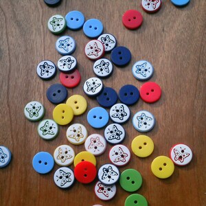 May include: Assortment of colourful buttons with butterfly designs. The buttons are in various colours, including red, blue, yellow, green, and white. The buttons are arranged on a wooden surface.