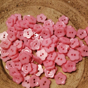 Op de afbeelding: Een stapel roze bloemvormige knopen met elk twee gaten. De knopen zijn willekeurig gerangschikt op een bruin houten oppervlak.