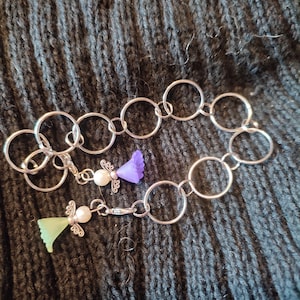Könnte beinhalten: Silberfarbenes Kettenarmband mit einer Reihe von verbundenen Kreisen. Das Armband hat zwei Anhänger: einen mit einer lila Blume und einer Perle und einen weiteren mit einem grünen Engel und einer Perle. Das Armband wird auf einer dunkelgrauen Strickoberfläche präsentiert.