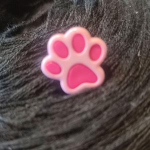 Könnte beinhalten: Ein rosa Pfotenabdruck-Anhänger mit einem helleren rosa Rand. Der Anhänger ist ein Accessoire zum Thema Haustiere, wahrscheinlich für ein Armband oder anderen Schmuck. Der Anhänger ist vor einem dunklen, strukturierten Hintergrund platziert.