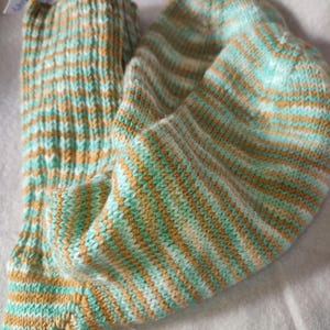 Op de afbeelding: Een paar handgebreide sokken met horizontale strepen in tinten turquoise, bruin en crème. De sokken zijn opgevouwen en tonen het getextureerde breipatroon. Een klein label is aan één sok bevestigd.