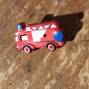 Può includere: Un camion dei pompieri giocattolo rosso con dettagli blu sulla parte superiore e posteriore. Il giocattolo ha ruote nere e dettagli bianchi. Il camion dei pompieri è su una superficie di legno.