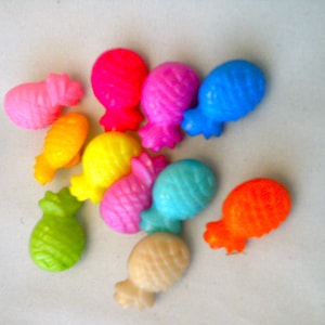 Könnte beinhalten: Eine Sammlung bunter, plastikartiger, fruchtförmiger Perlen. Die Perlen zeigen eine Vielzahl von Früchten wie Ananas, Bananen und andere tropische Früchte in Pink-, Gelb-, Grün-, Blau- und Orangetönen. Die Perlen liegen auf einer weißen Oberfläche.