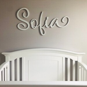 May include: White wooden name sign with the name "Sofia" written in a cursive script. The sign is hanging on a light beige wall above a white crib.