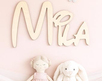 May include: A wooden sign spelling "Mia" in cursive script, mounted on a pink wall, with three decorative butterflies. A fabric doll in a pink dress and a white plush bunny sit below the sign, creating a nursery theme.