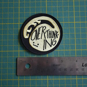 May include: A black and white embroidered patch with a white outline of a creature and the text "OVER THINKING" in white.