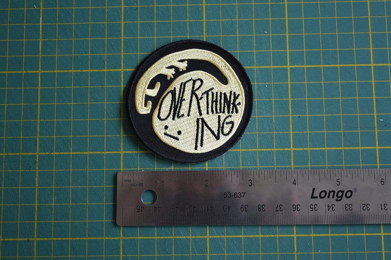 May include: A black and white embroidered patch with a white outline of a creature and the text "OVER THINKING" in white.