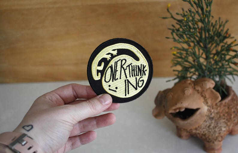 May include: Black and gold embroidered patch with the text "Overthinking" in a stylised font.