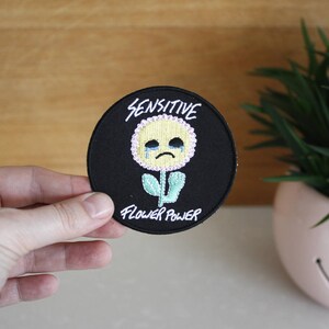 May include: Black and white embroidered patch with a yellow flower with a crying face. The text "Sensitive Flower Power" is written around the flower.