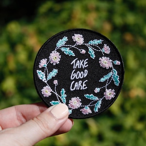Könnte beinhalten: Ein schwarzer gestickter Aufnäher mit einem floralen Kranzdesign und dem Text "Take good care".