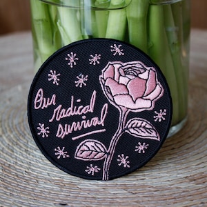 May include: A black embroidered patch with a pink rose and the text "Our radical survival" in pink cursive lettering. The patch is surrounded by small pink snowflakes.