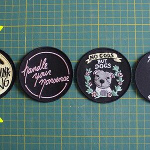 May include: Four round embroidered patches with different designs. The first patch is black with gold embroidery and says "Overthinking". The second patch is black with pink embroidery and says "Handle your nonsense". The third patch is black with white and green embroidery and says "No Gods But Dogs". The fourth patch is black with yellow embroidery and says "Sensitive Flower Power".