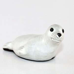 May include: A small, light gray seal figurine with black eyes and a black nose. The seal is lying down, with its flippers tucked in. The figurine is made of a smooth material and has a detailed face. The seal measures approximately 4 inches long.