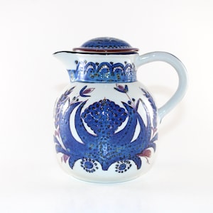 Puede incluir: Una jarra de cerámica con tapa, con una base blanca adornada con patrones y acentos florales azules. La jarra tiene un asa curva y un pico. La tapa es de un azul más oscuro con un borde marrón. El diseño general es de estilo vintage.