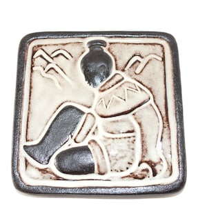 May include: Square ceramic tile with a relief design of a seated figure, mountains, and a decorative border. The figure is depicted in black and cream colors, with a dark brown border. The tile is likely for decorative purposes.