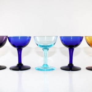 Puede incluir: Cinco copas de champán de colores en fila. Las copas tienen un cuenco redondeado y un tallo. Los colores son morado, azul, azul claro, azul y ámbar. Los tallos y las bases son oscuros.