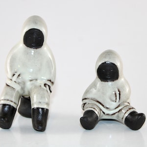 May include: Two ceramic Inuit figures. The figures are wearing white hooded parkas with black faces and boots. One figure is seated with legs extended, and the other is seated with legs bent. The figures have a glossy finish.