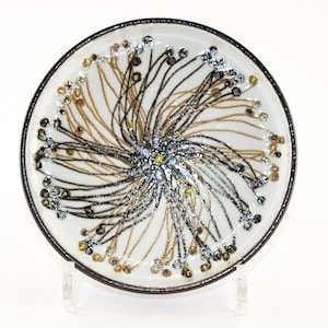 Puede incluir: Un plato de cerámica blanco con borde negro y un diseño central de líneas radiales en negro, dorado y plateado. Las líneas tienen pequeños adornos redondos. El plato se exhibe sobre un soporte transparente.