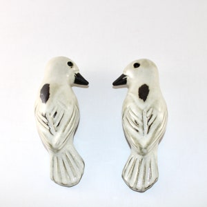 May include: Two ceramic bird figurines. Each bird is white with a black beak and a dark spot on the side. The birds are facing each other, and the background is plain white.